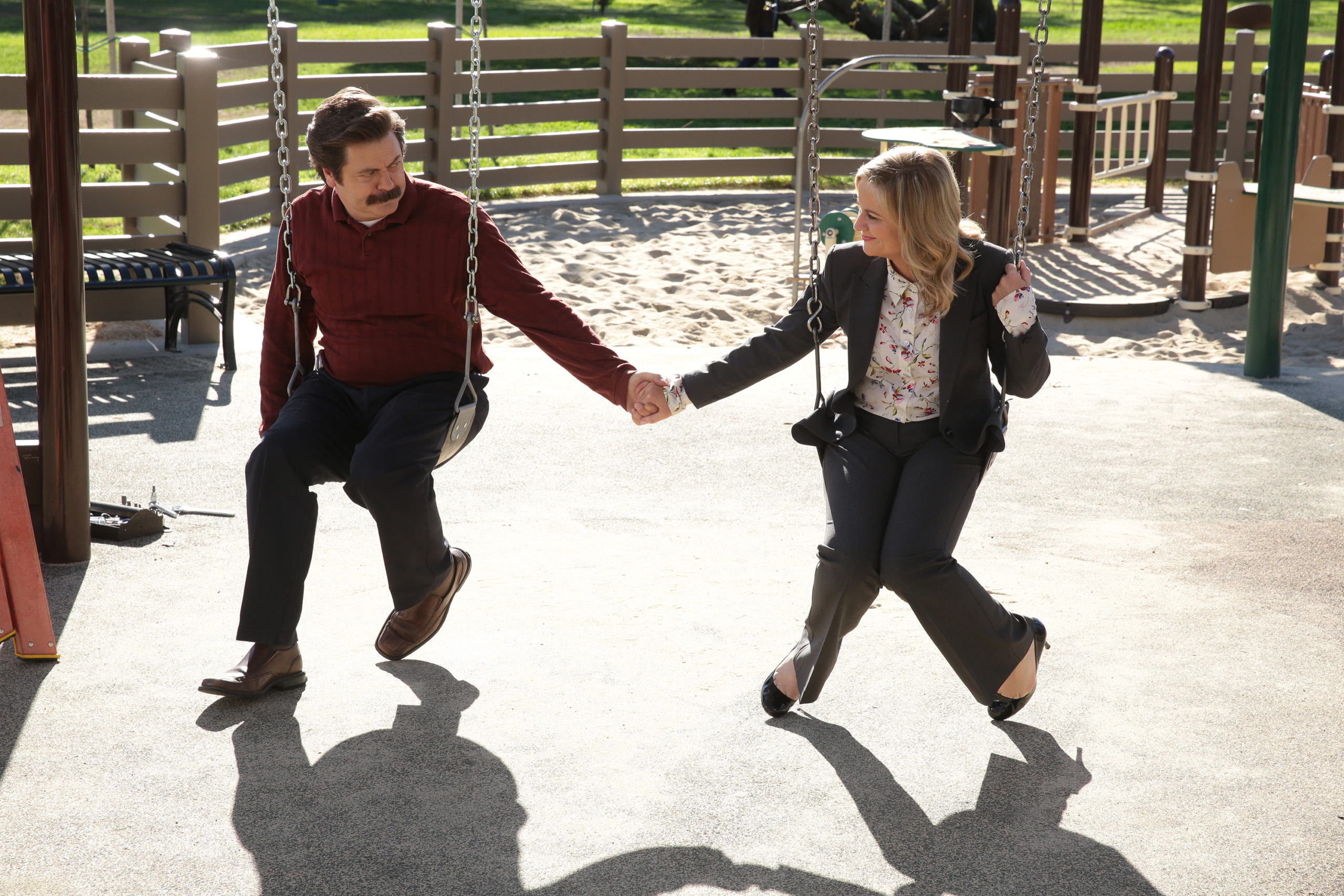 Nick Offerman as Ron Swanson and Amy Poehler as Leslie Knope holding hands on a swing in Parks and Recreation - 'One Last Ride'