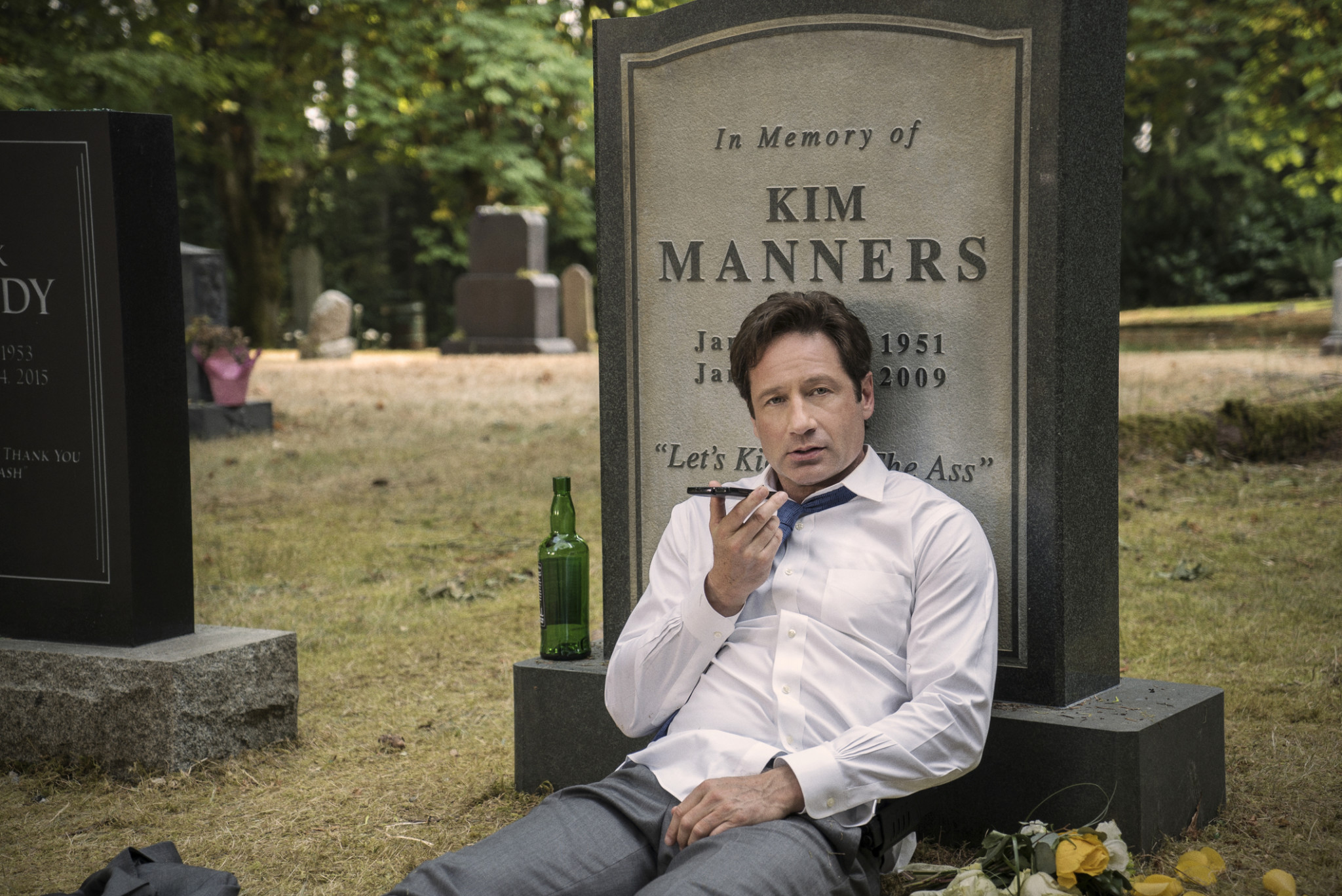 David Duchovny talking on his phone in front of the gravestone of Kim Manners in The X-Files