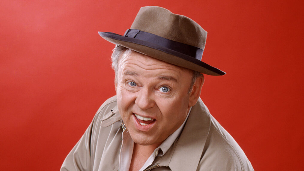 Carroll O'Connor (1924-2001), US actor, wearing a brown hat with a dark blue band and a beige jacket, and holding a cigar, in a studio portrait, against a red background, issued as publicity for the US television series, 'All in the Family', USA, circa 1975. The sitcom starred O'Connor as 'Archie Bunker'. (Photo by Silver Screen Collection/Getty Images)