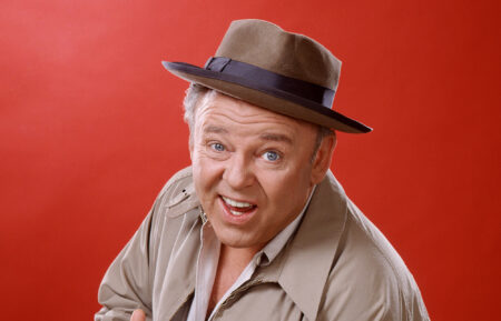 Carroll O'Connor (1924-2001), US actor, wearing a brown hat with a dark blue band and a beige jacket, and holding a cigar, in a studio portrait, against a red background, issued as publicity for the US television series, 'All in the Family', USA, circa 1975. The sitcom starred O'Connor as 'Archie Bunker'. (Photo by Silver Screen Collection/Getty Images)