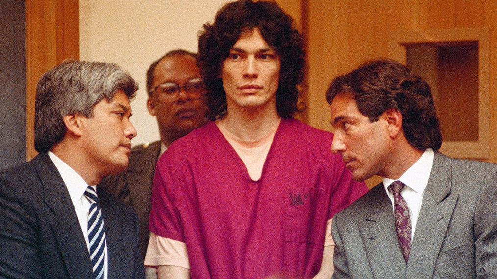 A still from Netflix documentary Night Stalker: The Hunt for a Serial Killer. Richard Ramirez, wearing fuschia prison scrubs, stands up in court, flanked by officials in grey suits and ties.