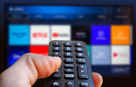A man is holding a remote control of a smart TV in his hand. In the background you can see the television screen with streaming entertainment apps for video on demand