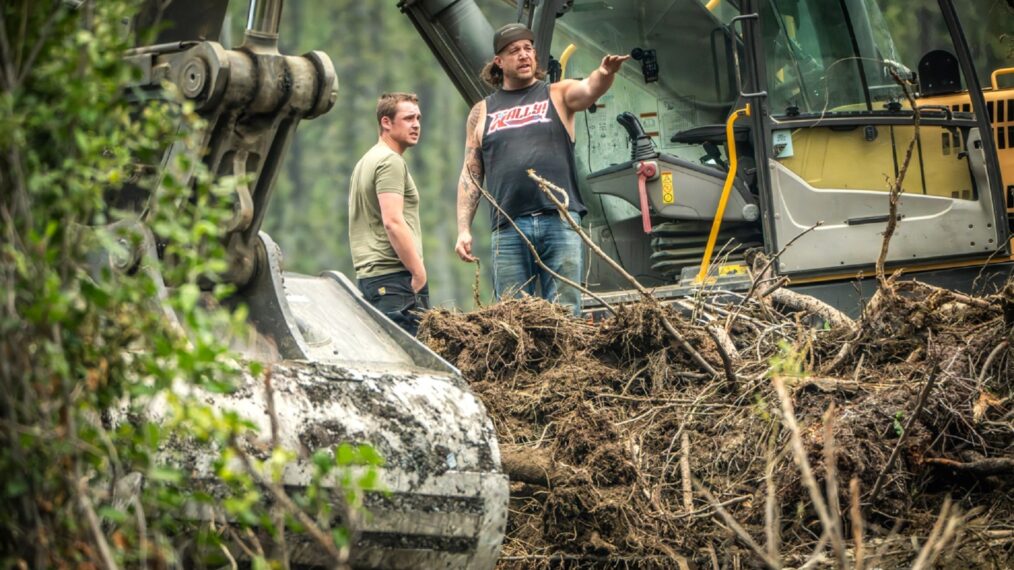 Rick Ness and Bailey Carten mid conversation by excavator, busy foreground in Gold Rush