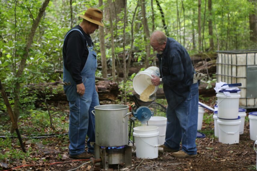 Eric "Digger" Manes and Mark Ramsey filling a pot with corn in the woods.