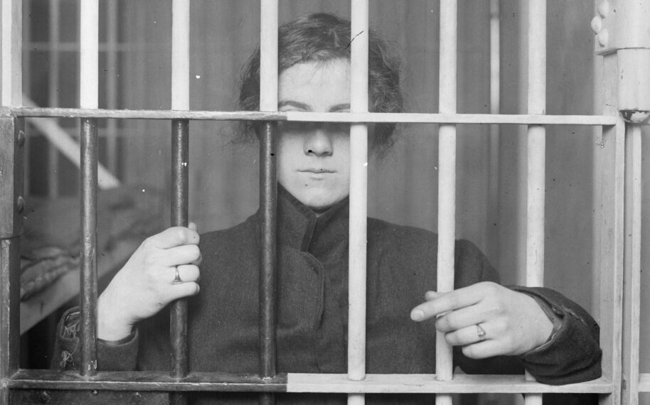 Crime and Punishment; An unidentified, young woman stands behind the bars of a jail cell in Denver, Colorado. She wears a wool coat with a raised collar and two rings on her hands.Credit: Denver Public Library