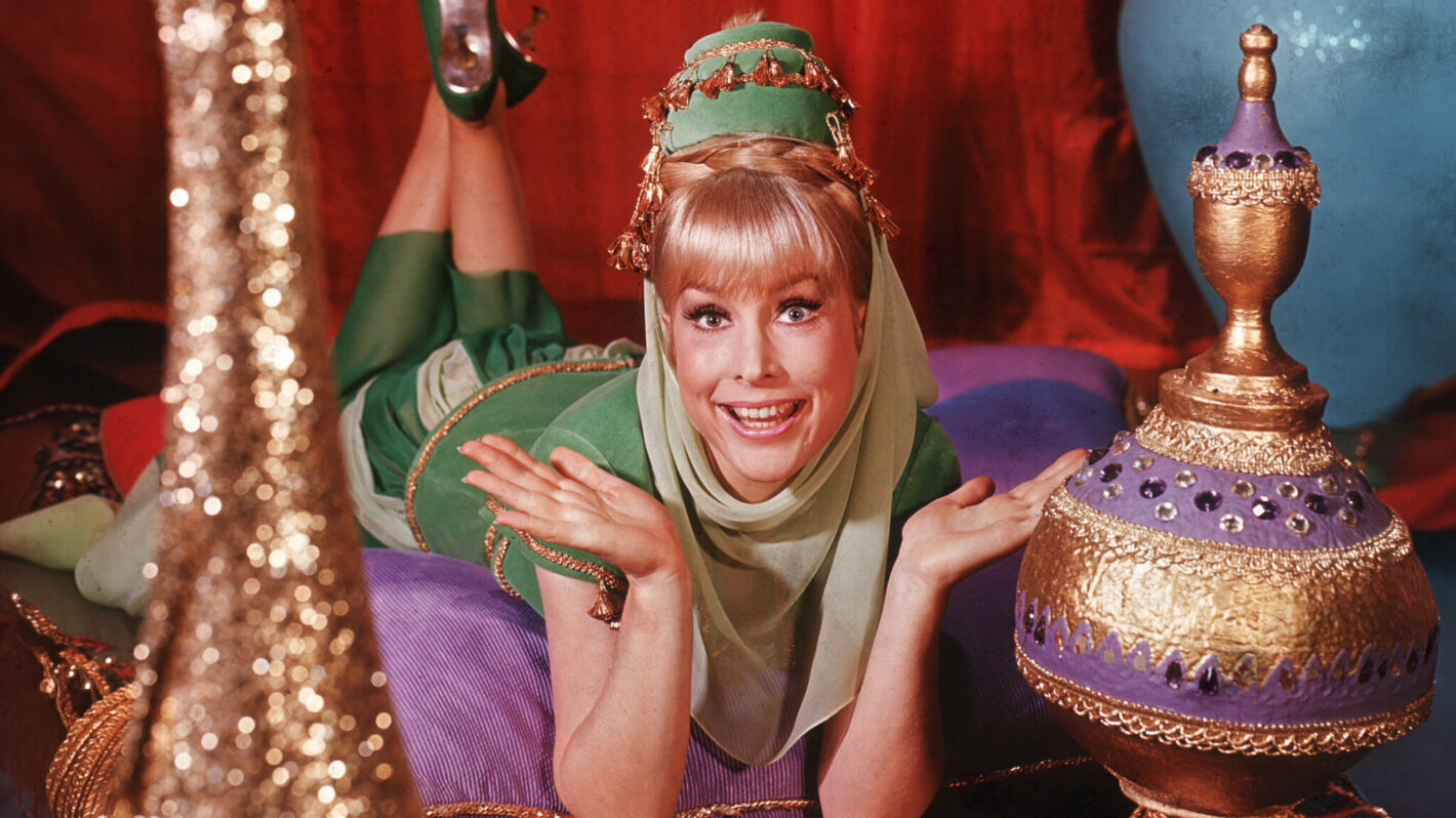 1965: American actor Barbara Eden holds her hands up and smiles while lying on her stomach on top of cushions on an oriental rug, in a promotional portrait for the television series 'I Dream of Jeannie'. Eden is dressed in a green genie costume. Colorful vases surround her.