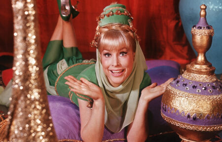 1965: American actor Barbara Eden holds her hands up and smiles while lying on her stomach on top of cushions on an oriental rug, in a promotional portrait for the television series 'I Dream of Jeannie'. Eden is dressed in a green genie costume. Colorful vases surround her.
